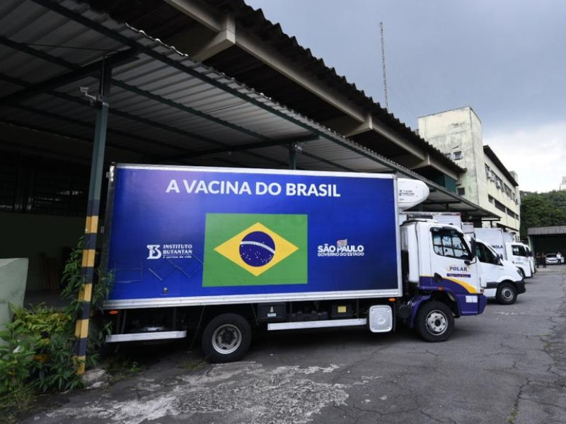 O caminhão com as doses está estacionado. Na laterial do caminhão, em azul, está escrito A Vacina do Brasil. Do lado esquerdo tem o logo do Instituto Butantan, ao centro uma bandeira do Brasil, e do lado direito o emblema do Governo do Estado de São Paulo